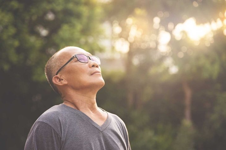 Happy Asian senior male wear glasses relaxing and breathing fresh air at park with sunlight and copy space. Elderly male smiling standing over nature on holiday. Carefree, Lifestyle, Wellness.